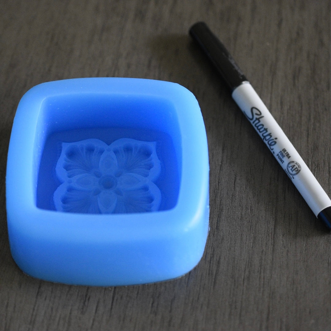 A square soap mold with a floral design rests next to a sharpie pen on a wooden surface for size comparison. The sharpie pen is longer than the mold in length. The mold is blue.
