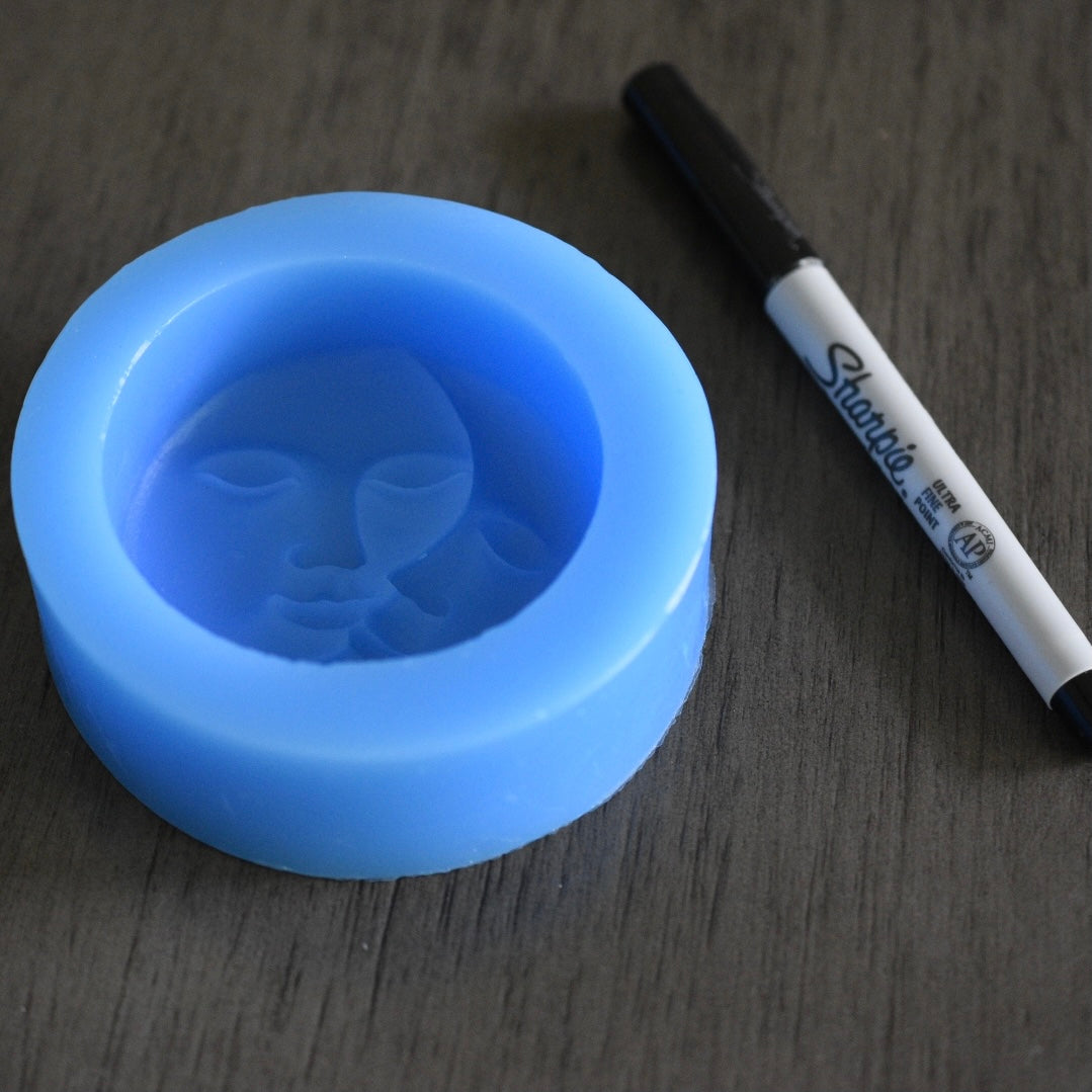 A round sun and moon soap mold rests next to a sharpie pen on a wooden surface for size comparison. The sharpie pen is slightly longer than the mold. The sun and moon are facing each other with stoic faces. The mold is blue.