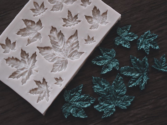 An assortment of maple leaves made from soap next to a small maple leaf mold on a wooden surface. The soap maple leaves are colored green. The mold is grey.