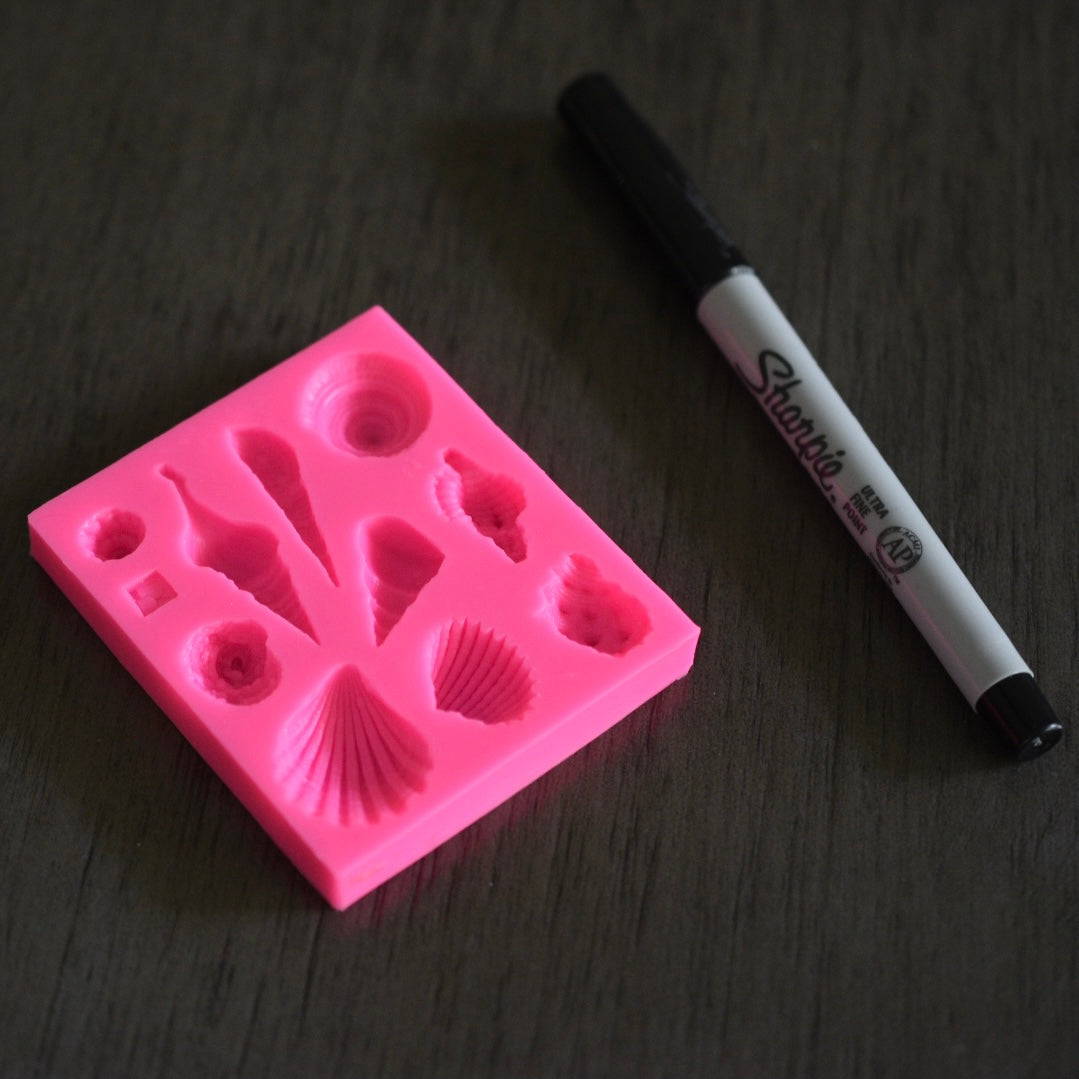 A top down view of a square soap mold with seashell designs rests next to a sharpie pen on a wooden surface for size comparison. The sharpie pen is slightly longer than the square mold. The mold is pink.