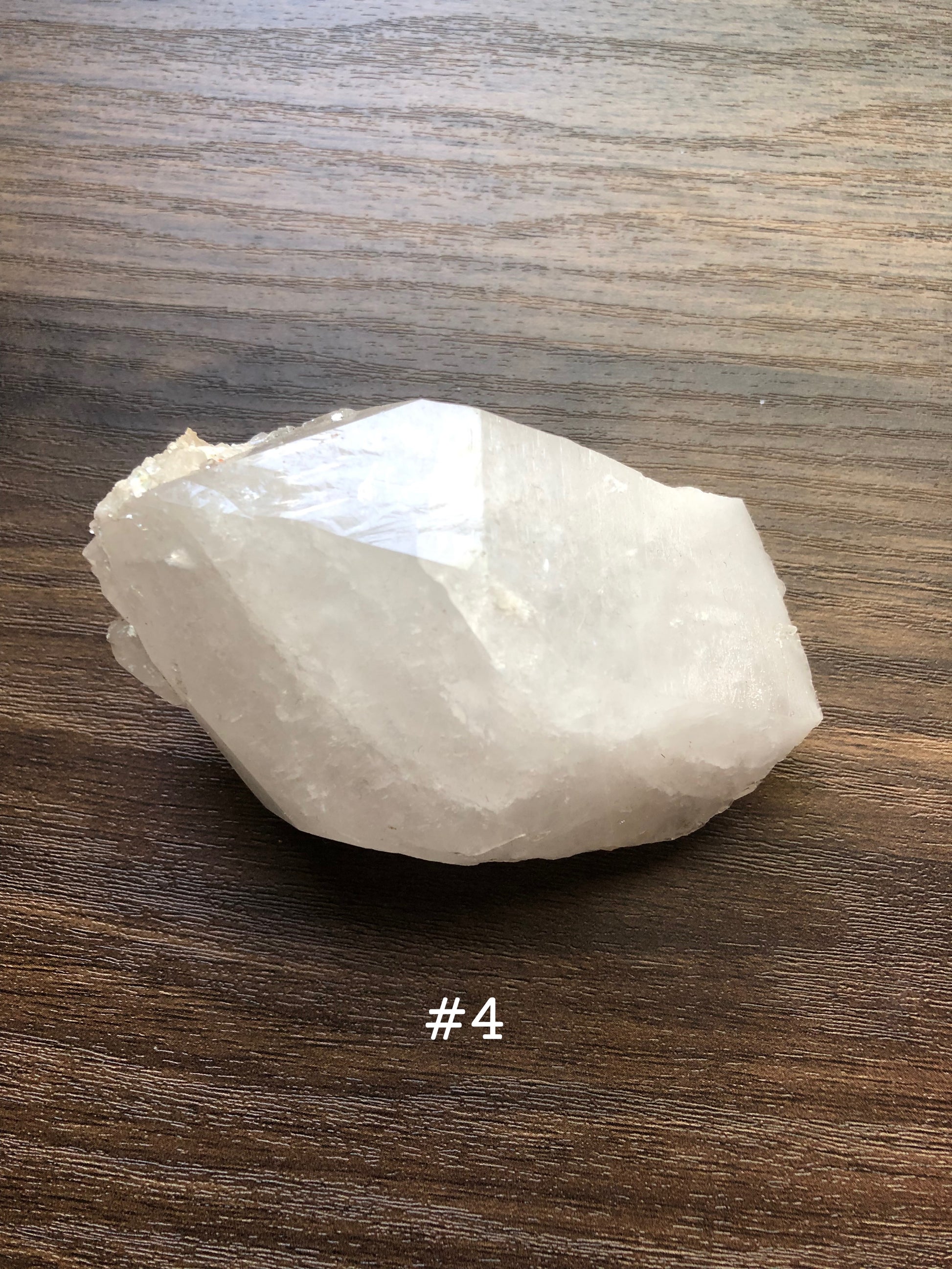 A large, rough cut quartz crystal rests on a wooden surface. It is wide and circular in shape. The crystal is relatively clear. The number 4 is shown on the picture.
