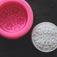 A round bar of soap with an elegant, filigree design rests next to round soap mold on a wooden surface. The mold is pink.