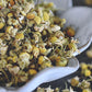 A close up view of an assortment of chamomile buds with short stems attached. The chamomile buds are a soft yellow with a light green stem. The chamomile rests in a white, flower shaped dish on a wooden surface. Chamomile buds are scattered about in the background.