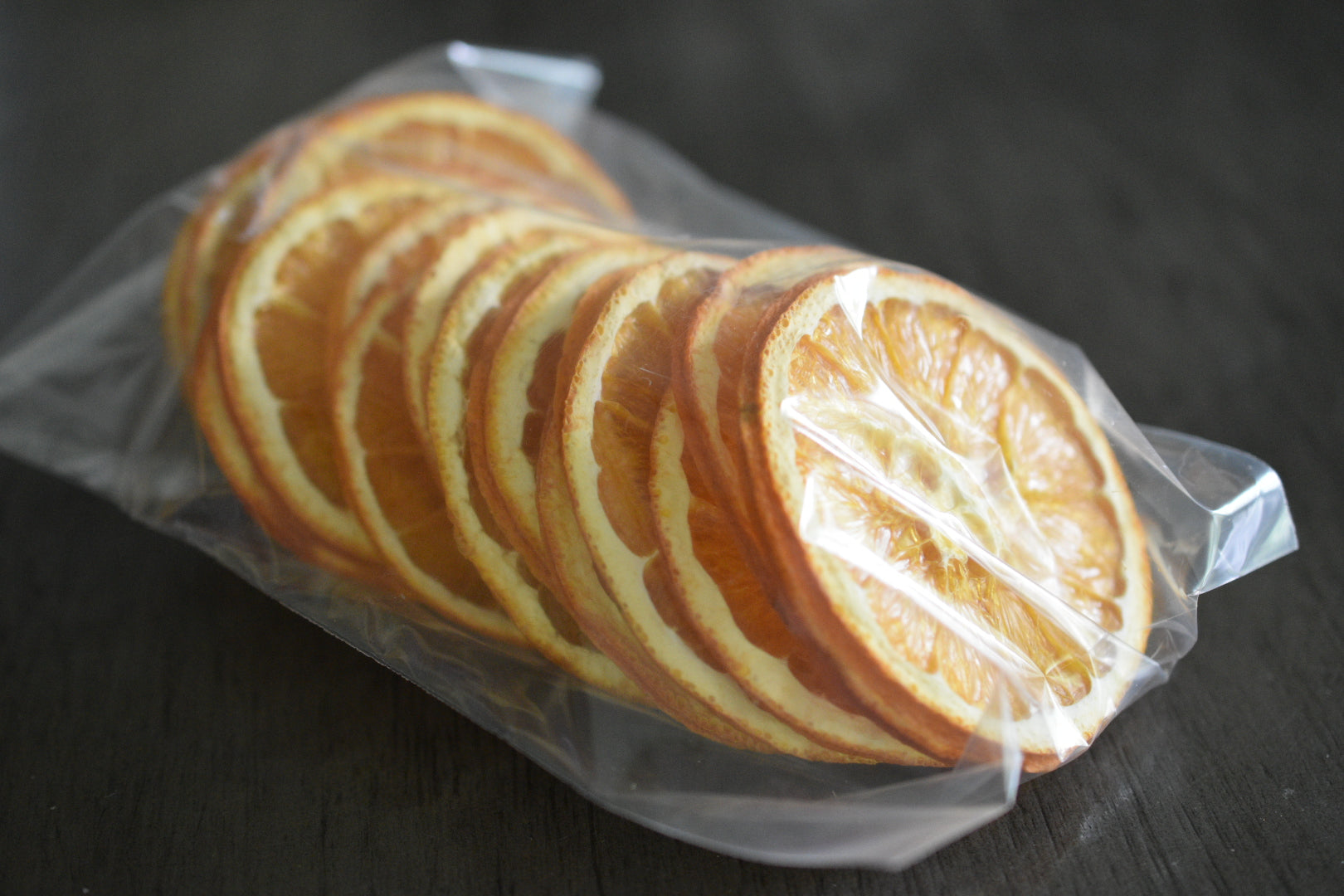 A plastic bag filled with dried orange slices rests on a wooden surface to show the packaged product. The orange slices are deep orange in color.