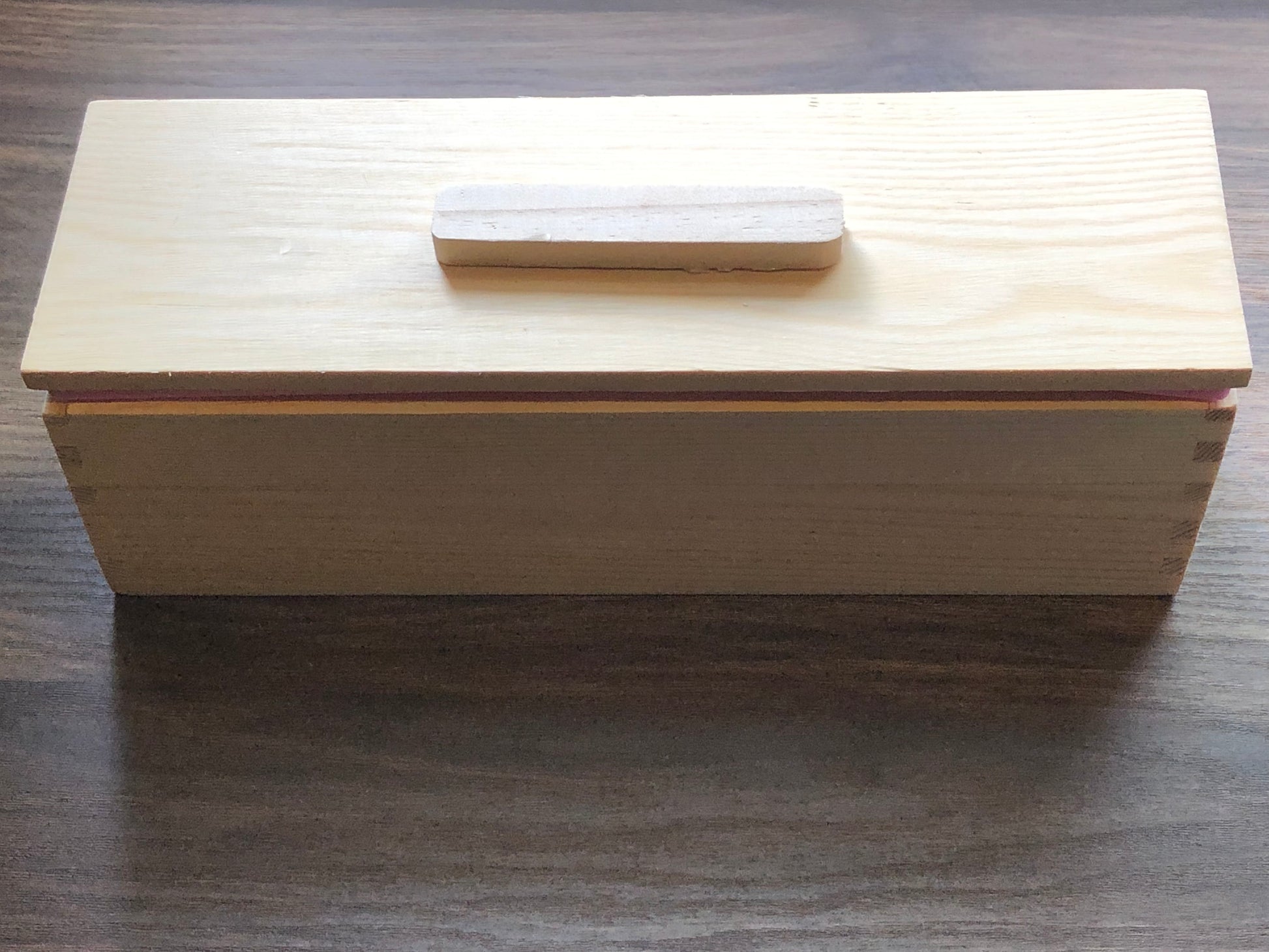 A loaf shaped wood box that has a loose lid with a small handle on the top of the lid rests on a wooden surface.  The wood of the mold is light in color.