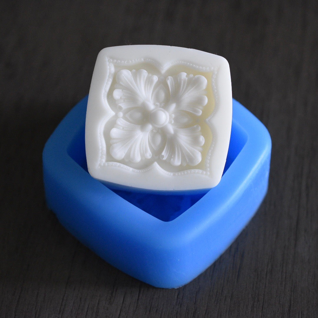 A square soap bar with a floral design rests in a square mold on a wooden surface. The soap bar is white. The mold is blue.