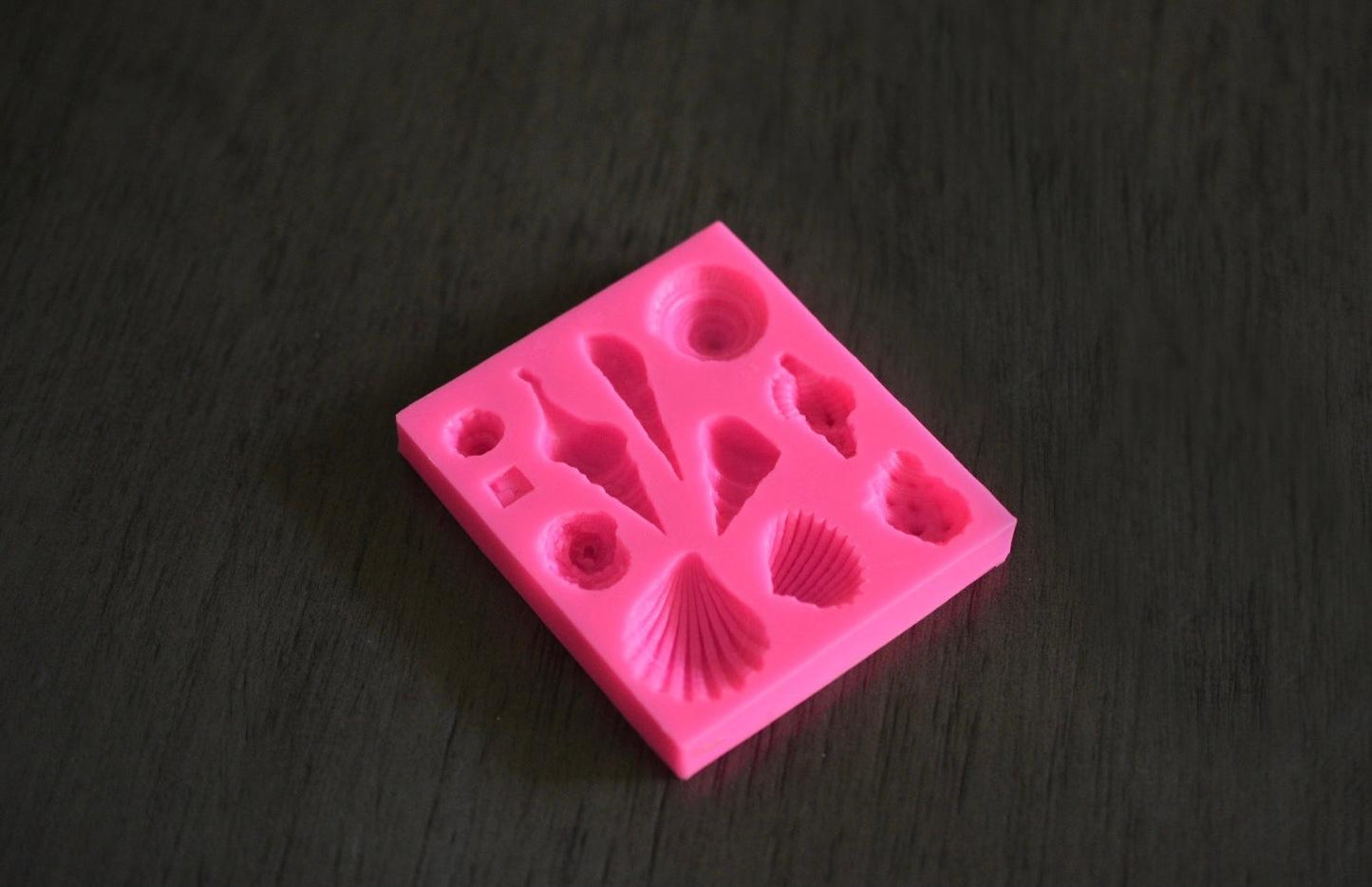 A top down view of a square soap mold with seashell designs rests on a wooden surface. The mold is pink.