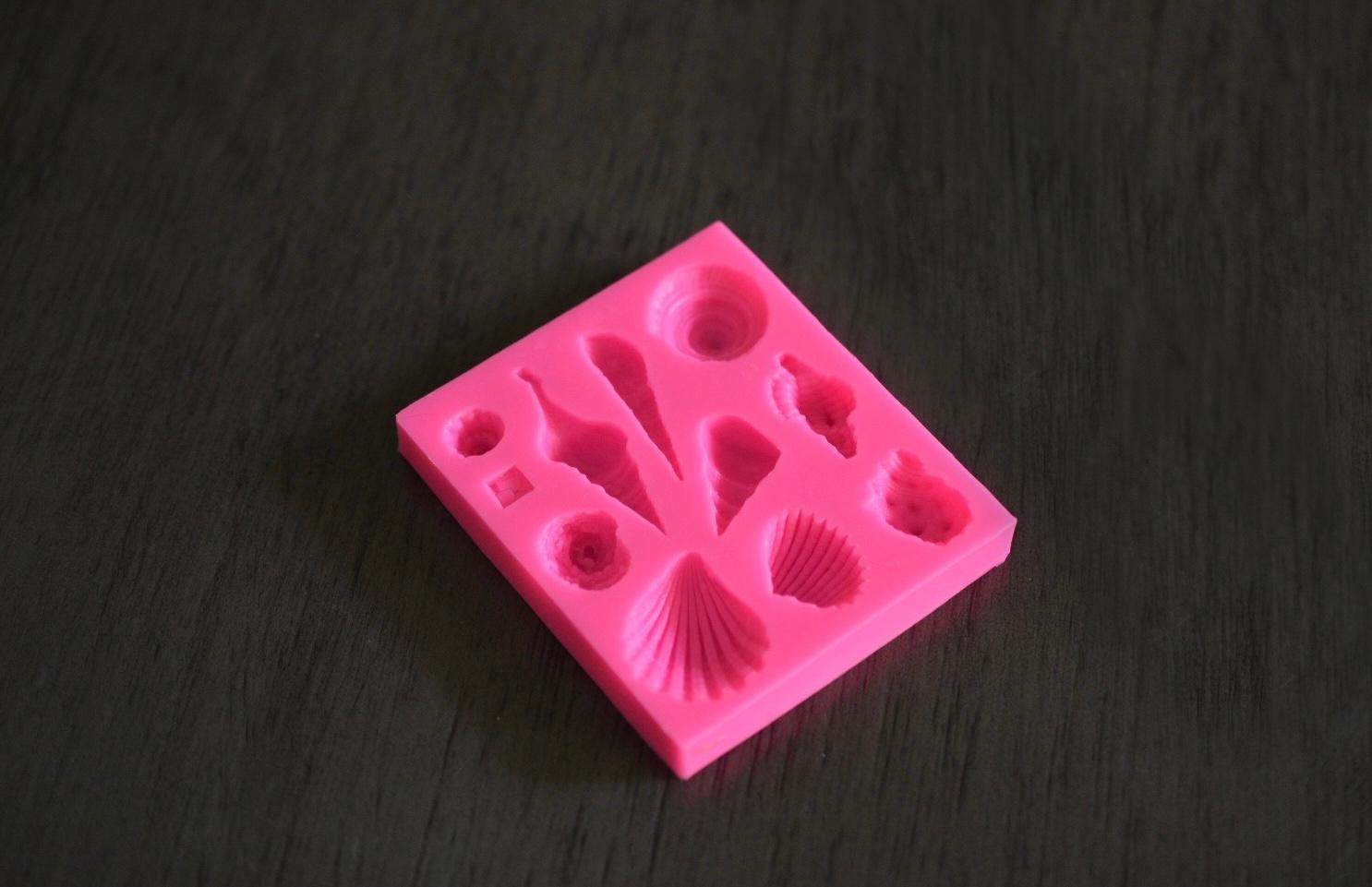 A top down view of a square soap mold with seashell designs rests on a wooden surface. The mold is pink.