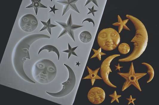 An assortment of moons and stars made of soap sits next to a rectangle mold. The moons and stars have been colored gold. The moons have stoic faces on them. The mold is grey.